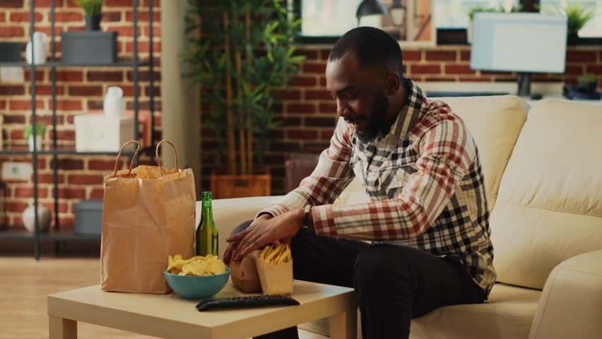 Smiling man biting on hamburger in living room, eating cheeseburger with fries and bottle of alcohol. Young adult watching television film and having dinner, feeling relaxed with takeaway meal.