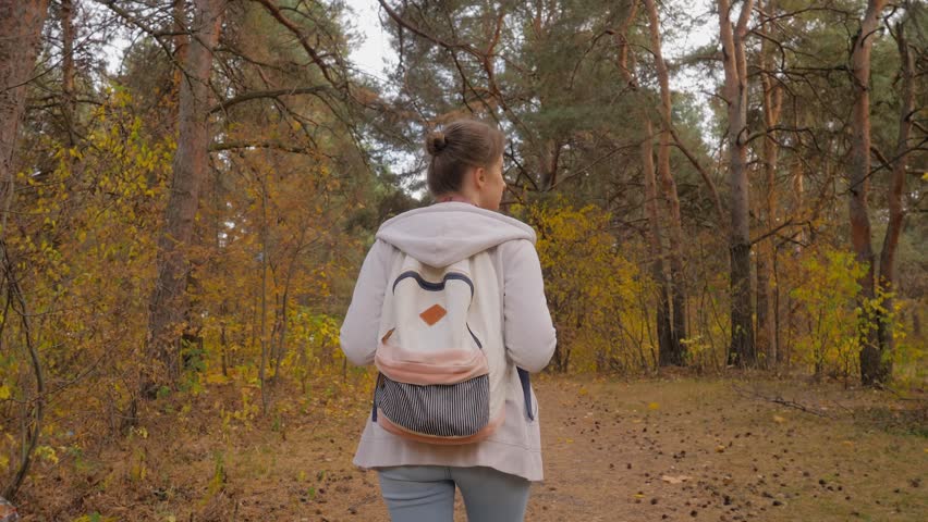 Back view of young woman with backpack walking in autumn park, forest - wide angle steadicam follow shot. Active outdoor lifestyle, leisure time, freedom and adventure concept