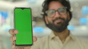 Focus on mobile phone with green screen holding by Indian man in glasses. Blurred Arabian businessman in eyeglasses bearded entrepreneur hold smartphone with mock up chroma key display for ads indoors