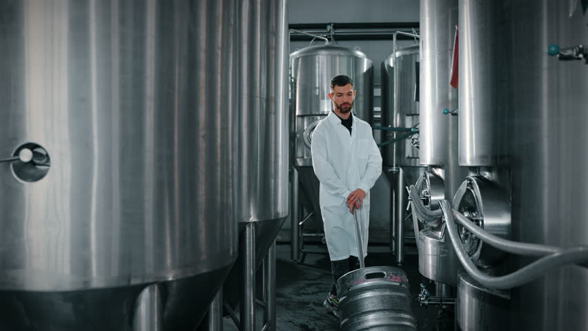 Modern brewery with large stainless steel tanks and worker with cart rolling a keg of beer. Food and beverages production facilities