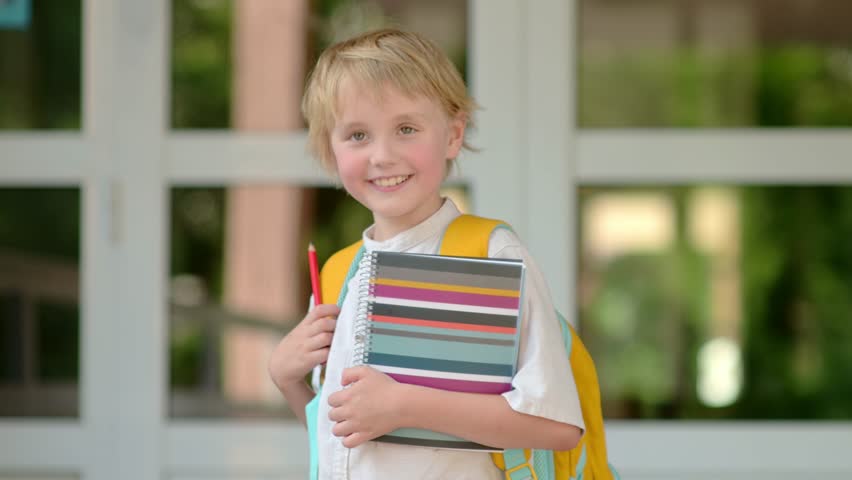 Slow motion portrait of happy school child. Cute schoolboy emotionally laughs and looks at you. Joyful boy before the start of the school year at the door of the school. Back to school Royalty-Free Stock Footage #1100375977