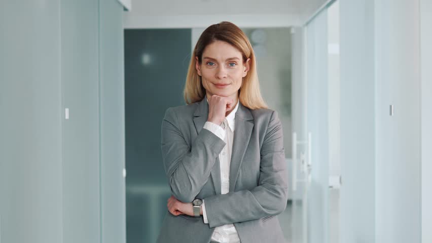 Middle aged confident focused business woman in formal suit standing in modern light office puts hand on chin smile looks at camera feels happy. 40s pretty positive female face portrait concept.  | Shutterstock HD Video #1100393715