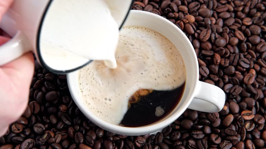 Steamed creamy milk poured into coffee cup to make cappuccino. Barista pouring milk to make latte. Footage video. Cafe business shop. Adding creamer into black coffee. Roasted coffee beans background. | Shutterstock HD Video #1100394443