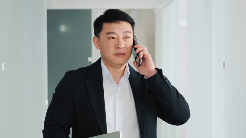 Chinese handsome businessman is discussing talking on mobile phone with a pleased smile standing in the office hall. Successful confident business man having friendly conversation, holding laptop.