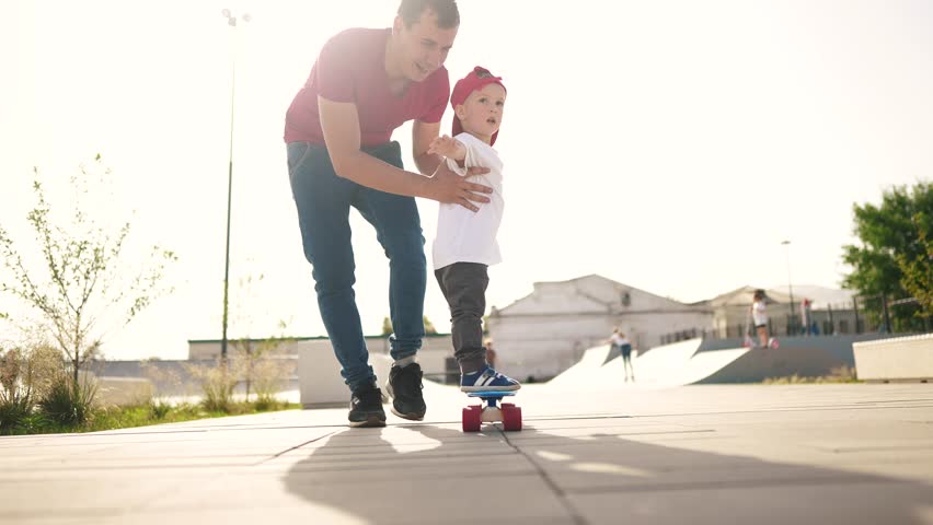 boy learn to skateboard. dad teaches son to ride a skateboard outdoors at the playground. father and boy play training concept. parent lifestyle teaching child son to skateboard Royalty-Free Stock Footage #1100396143