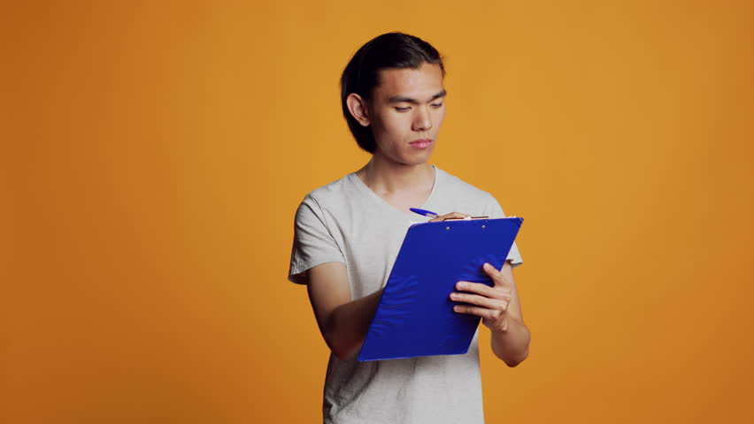 Confident young man writing and taking notes on files, using clipboard with papers to have informations. Carefree modern person holding pen to sign documents in studio, male model.