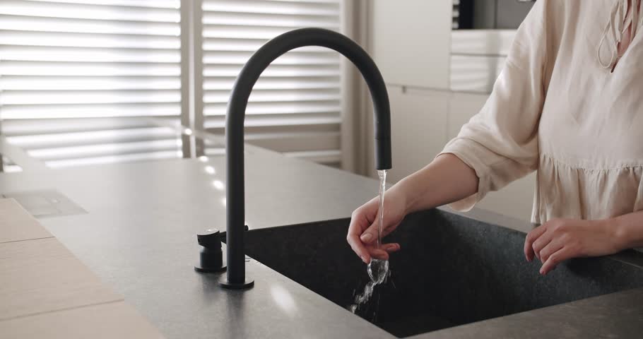 A woman turns on the tap in a modern white kitchen. Modern kitchen room in minimalist apartment, Black Kitchen sink and faucet, comfortable equipment. Royalty-Free Stock Footage #1100400841
