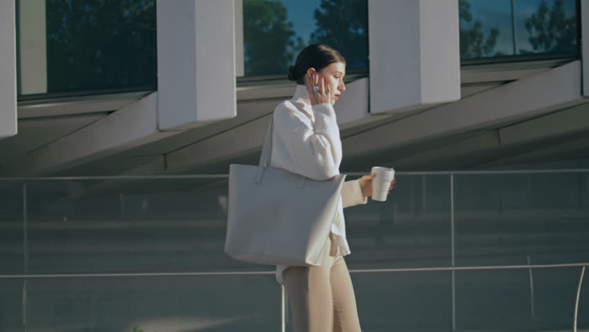 Busy trendy businesswoman talking through wireless headphones walking city street. Stylish confident business lady holding cup coffee takeaway going to office. Young woman calling using modern headset Royalty-Free Stock Footage #1100405141