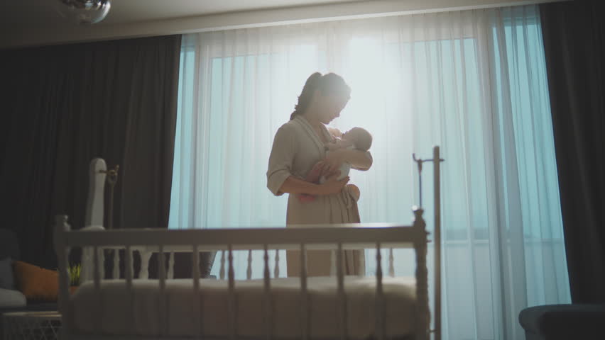 Young mother standing near large window, holding her newborn baby in arms and gently stroking her in slow-motion Royalty-Free Stock Footage #1100407477