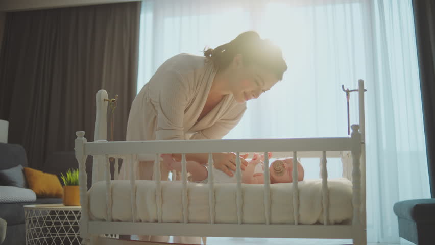 Beautiful smiling mother caring her little baby son lying in cradle and fixing pacifier position in baby's mouth | Shutterstock HD Video #1100407493