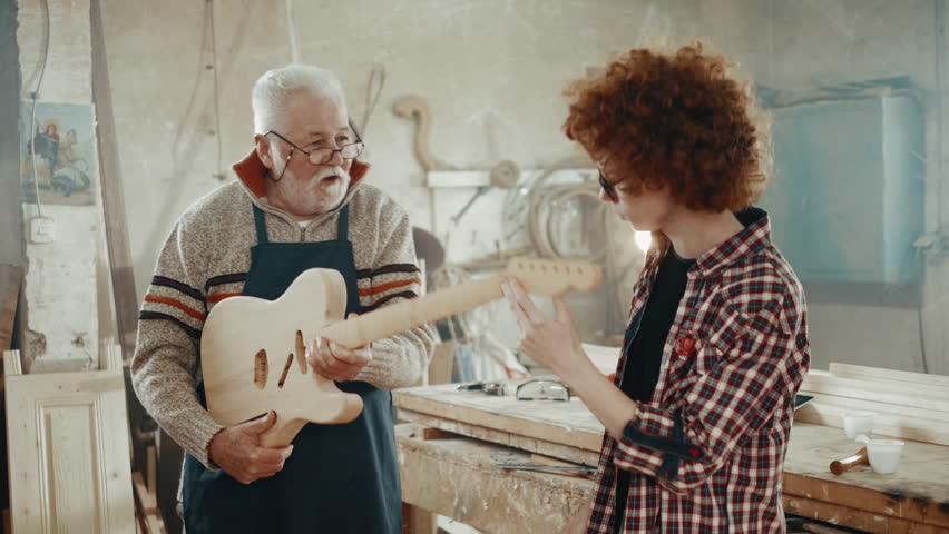 An old man and a teenager in a woodworking workshop high five a successfully made wooden guitar. Concept of carpentry Royalty-Free Stock Footage #1100409583