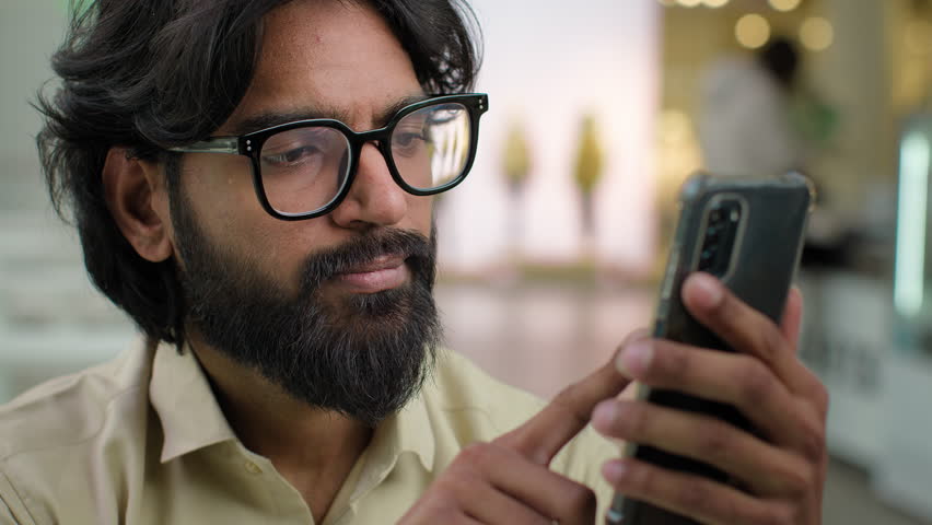 Indian male bearded man in glasses professional hold modern smartphone texting message in office indoors. Arabian businessman using mobile app browsing phone business chatting scrolling web online Royalty-Free Stock Footage #1100413779