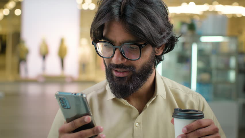 Male Indian bearded millennial professional hold modern smartphone drinking coffee in cafe. Arabian businessman in glasses man use mobile app for business look at phone drink tea at break in cafeteria Royalty-Free Stock Footage #1100413811