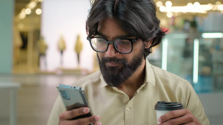 Male Indian bearded millennial professional hold modern smartphone drinking coffee in cafe. Arabian businessman in glasses man use mobile app for business look at phone drink tea at break in cafeteria | Shutterstock HD Video #1100413811