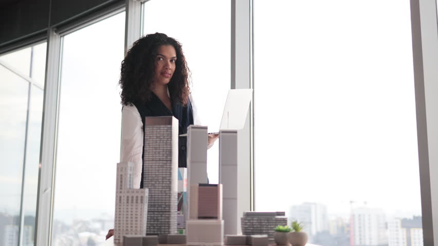 Smiling business woman real estate agents in stylish formal suit standing with laptop near 3d model skyscrapers of city architecture design at office with panoramic city view.