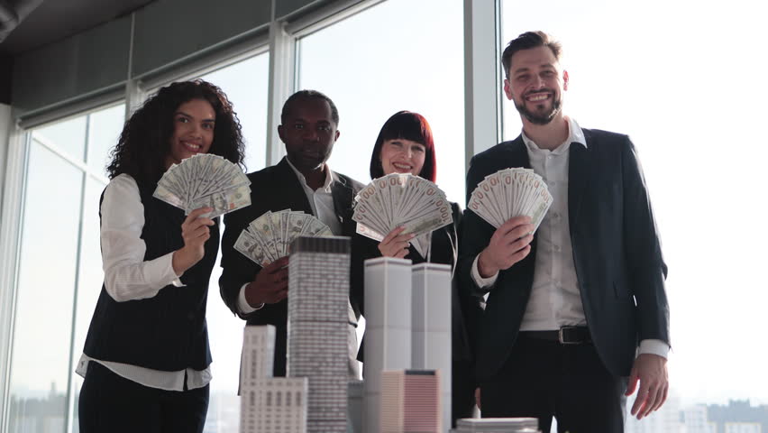 Team of diverse multiethnic business people holding bundle of american dollar cash money, staying near table with 3d skyscraper model at office interior with panoramic city view.
