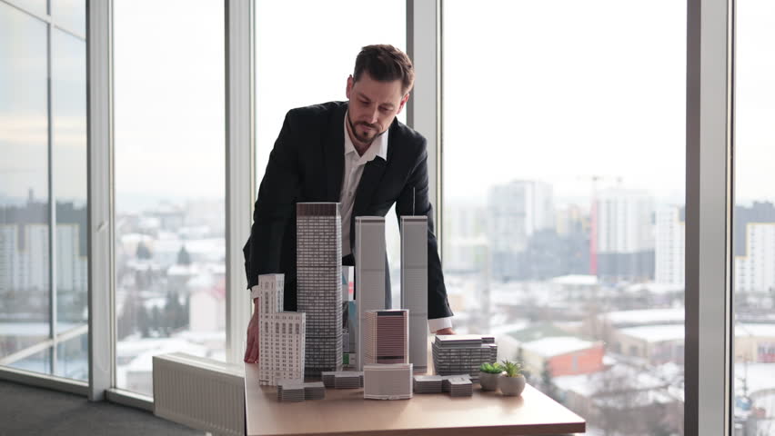 Indoor office portrait of bearded young business man, wearing white shirt and black suit, standing near wooden table with 3D megalopolis model in modern office interior with panoramic city view.