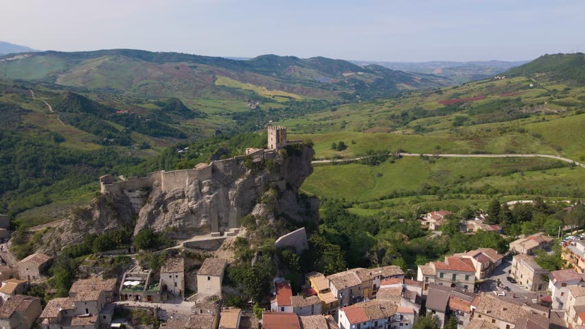 Drone shot flying towards a medieval castle on the side of a cliff revealing the small village below and the mountains. The castle is called Roccascalegna and is located in Abruzzo, Italy.