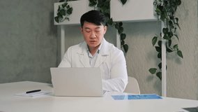 Asian male general practitioner give medical support to patient distantly use video call app indoor, look at computer screen talk to client makes distance speech. Remote medicine consultation concept.