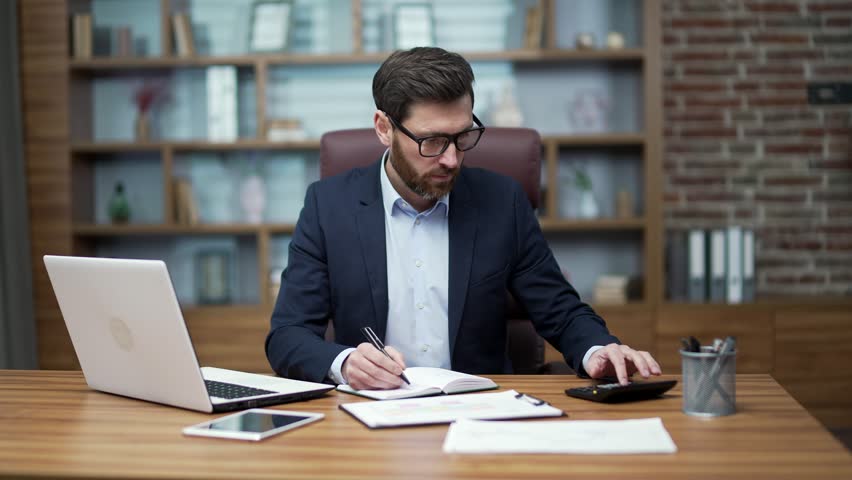 Concentrated busy business man financial analyst accountant bookkeeper or investor doing paperwork counting report graphs on calculator and make notes in working notebook at office workplace indoors | Shutterstock HD Video #1100419829