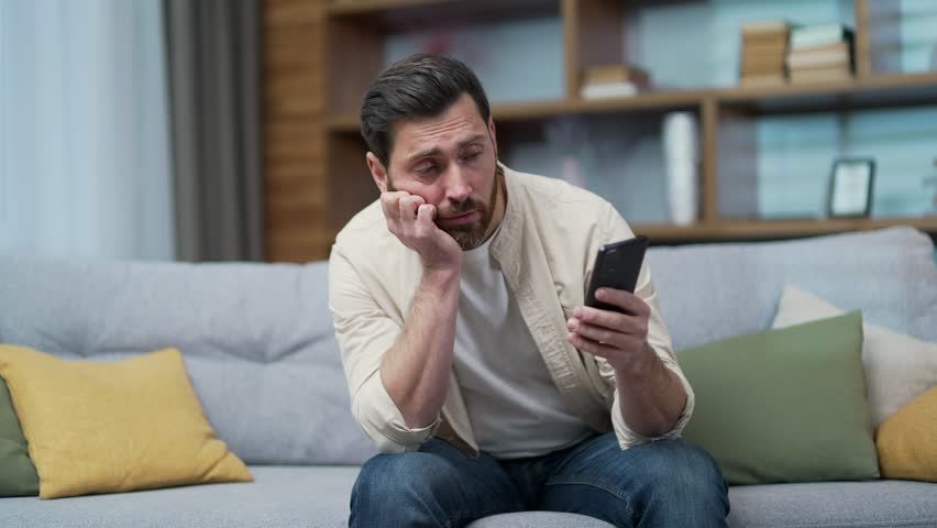 Boring tired bearded man scrolling feed of social networks products in internet store on smartphone sitting on couch in living room Exhausted sad male look at mobile screen without enthusiasm indoor Royalty-Free Stock Footage #1100419939