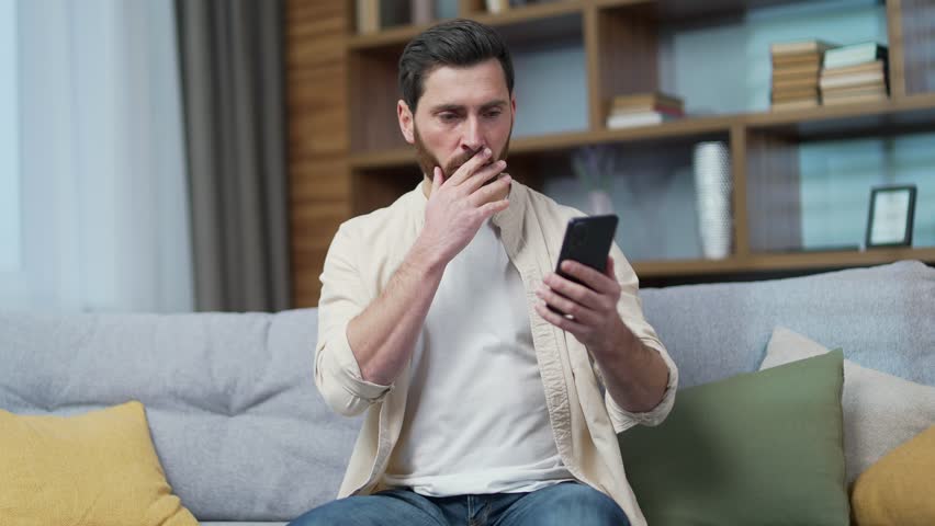 Shocked mature bearded man read bad news while received message email or browsing social media in smartphone at home Worried sad male stares at mobile phone screen with baffled puzzled face indoor  | Shutterstock HD Video #1100419941