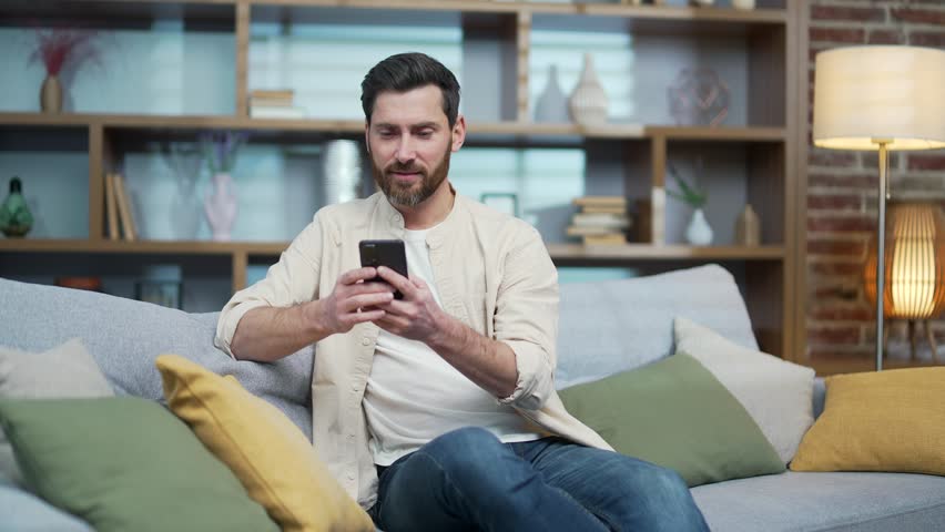 Amazed lucky man extremely happy after read message in mobile phone at home Excited bearded male looking at screen won pleased rejoices received great news on smartphone indoor alone Success Triumph Royalty-Free Stock Footage #1100419945