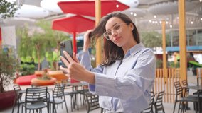 Woman taking selfie on smartphone camera outside. Brunet female person in glasses doing selfie photo on mobile phone camera on city street near outdoor cafe. Girl make a video call