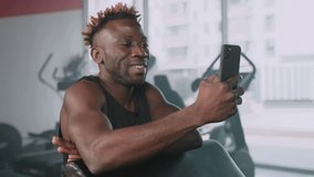 young black athletic man is viewing funny video in social networks during rest in gym workout, portrait of smiling african american guy with smartphone, use mobile phone in fitness center, new app