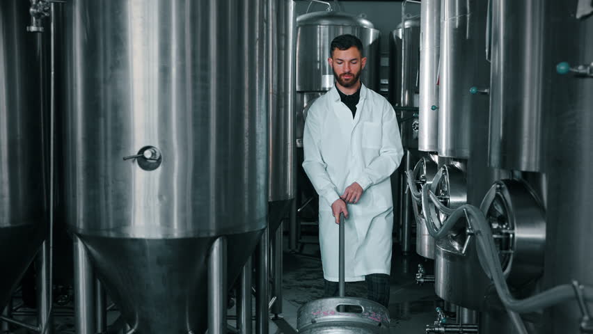brewer technologist is moving barrel with beer in workshop, between cylindrically-conical fermentation tanks, craft brewing factory, modern equipment