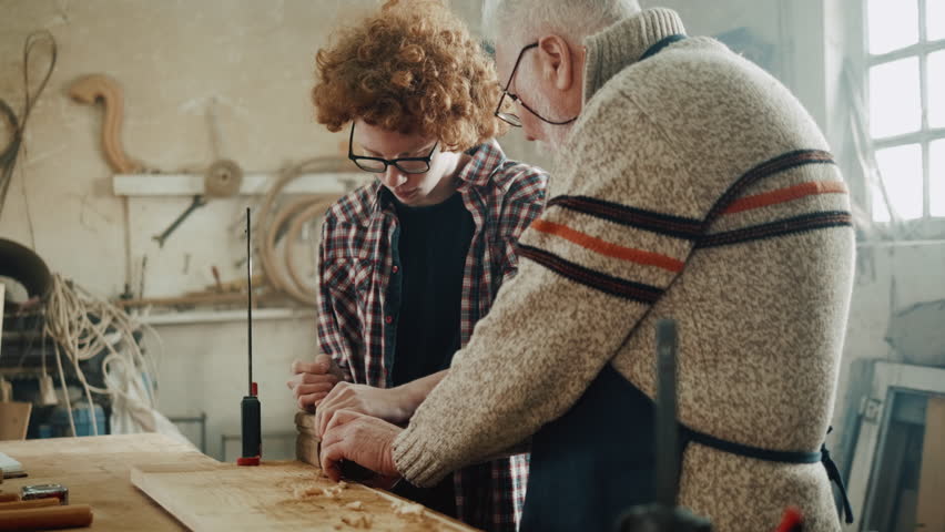 Old man teaching teen boy how to use a hand plane to square a board in his shop in the workshop Royalty-Free Stock Footage #1100424471