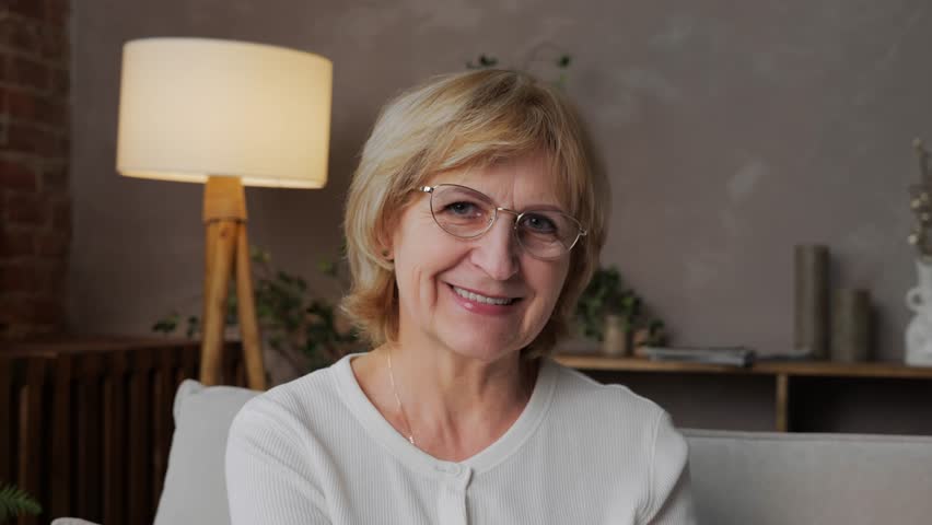 Portrait of a happy 60-year-old blonde woman with glasses in a home interior looking into the camera. Kind and positive and beautiful adult woman freelance programmer. High quality 4k footage Royalty-Free Stock Footage #1100425727