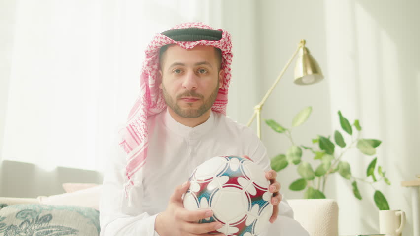 Middle eastern football fan supporting favorite team, watching match on TV. Young man holding soccer ball. Wearing traditional Islamic male clothes. Worship and culture concept.
