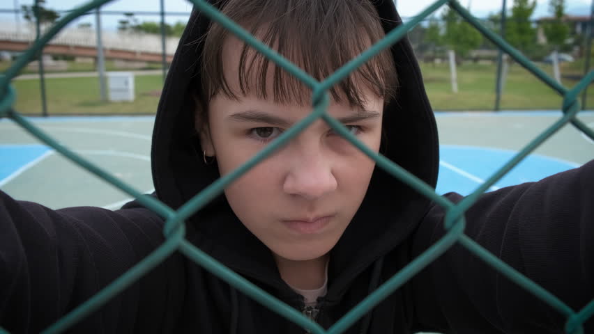 Adolescent behind the grid. A view of child hold the metal grid and look with angry on the school ground. Bullying at school.