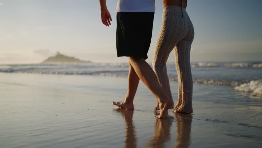Legs of happy couple holding hands and walking on the beach together enjoying summer side view. Feet of heerful boyfriend and girlfriend relaxing and taking a walk at the seaside at sunrise side shot. | Shutterstock HD Video #1100434921
