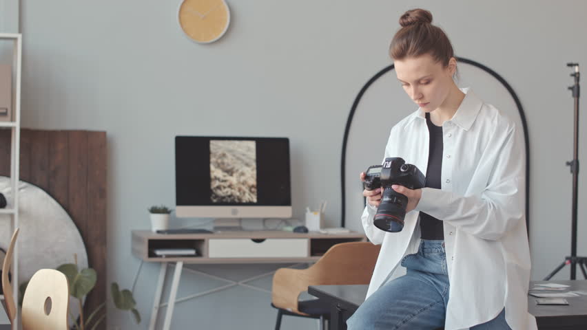 Medium slowmo of young Caucasian female photographer looking at photos on digital camera display while working in bright modern photo studio