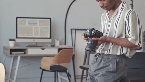 Medium slowmo of concentrated young African American male photographer looking at photos on digital camera display, working in photo studio Video Stok