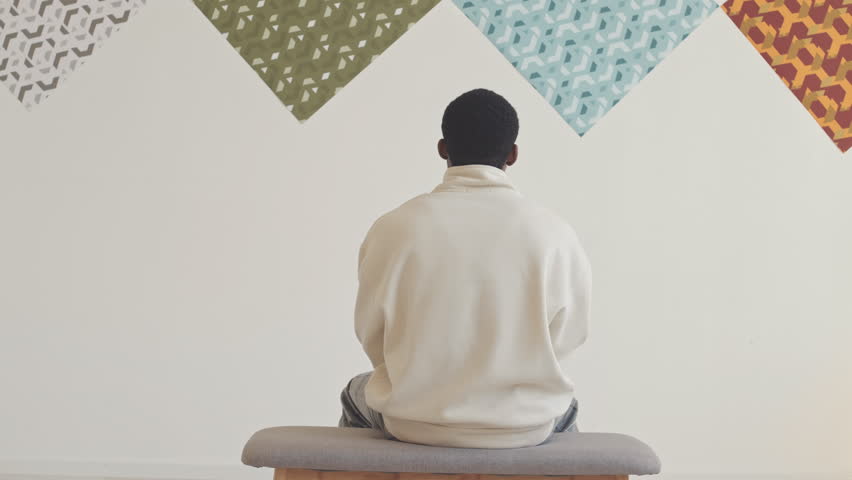 Back view of Black man looking at white wall with patterned pictures on it in modern art gallery