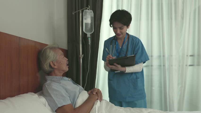 An elderly patient is lying in a hospital bed while a young doctor or nurse asks about their symptoms. The doctornurse listens attentively and provides medical advice to the patient. | Shutterstock HD Video #1100438987