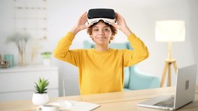 Young african girl at home put 3D glasses of virtual reality, sits at desktop near laptop. Amazing smiles, controls video game with hand movements and gestures, touching air. New technology concept.