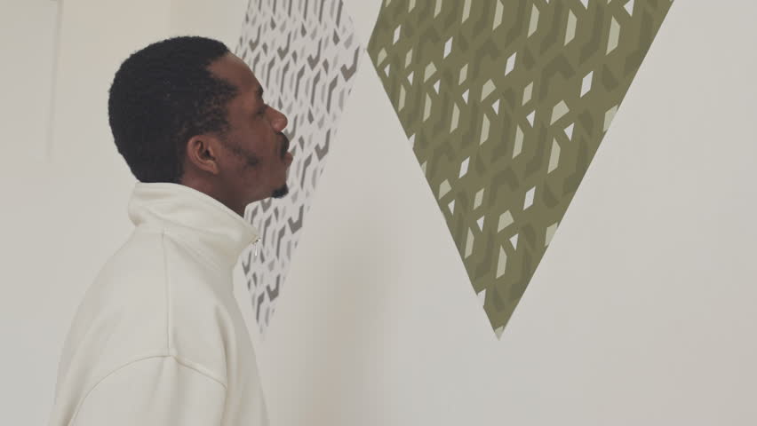 African American man standing close to minimalist pattern pictures on white wall in modern art gallery examining them