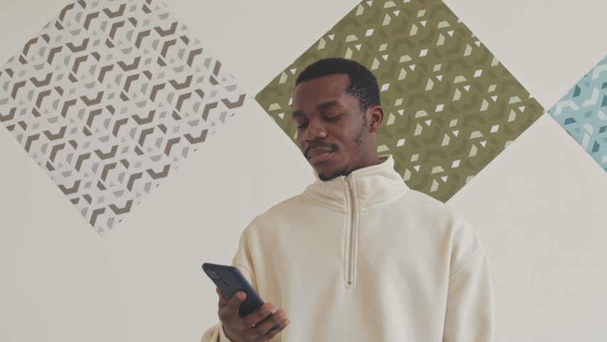 Young African American man standing against white wall with abstract pictures hanging on it and taking video on smartphone of unusual art objects, visiting modern art gallery
