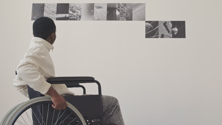 Slowmo of young African American man in wheelchair looking at pictures on walls in minimalist photo gallery