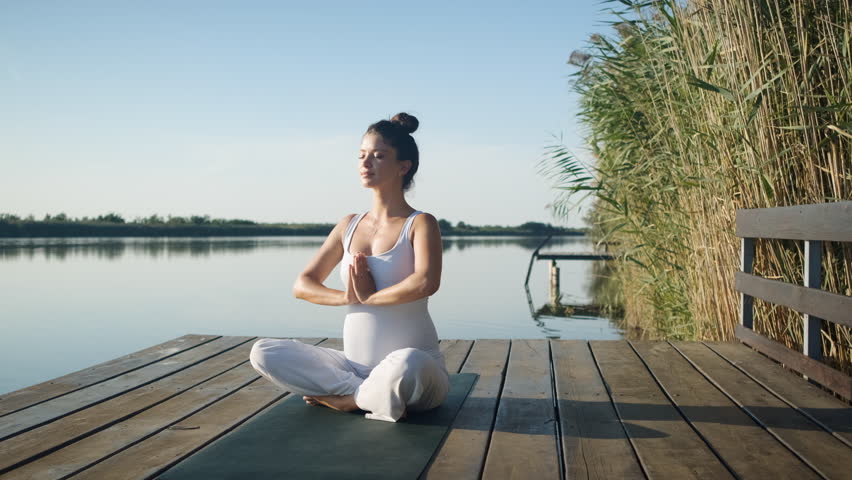 Pregnant woman doing yoga at lake during the day. Royalty-Free Stock Footage #1100442515