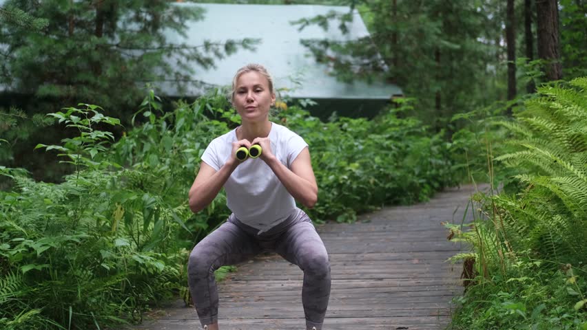 Pretty sportswoman doing squats with dumbbells outdoors in the forest, during a summer outdoor recreation