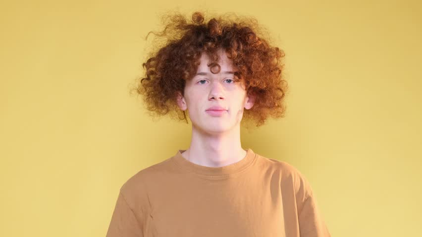 Curly red-haired calm young man shows the gesture of silence on a light brown background.