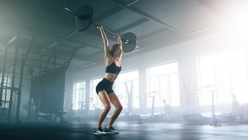 Portrait of a CrossFit sportswoman performing deadlift exercise in gym studio. Close-up shot of blonde-haired female bodybuilder lifting weights overhead. High quality 4k footage