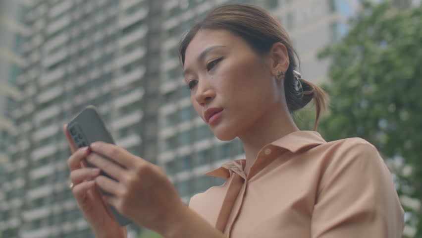Low angle chest up shot of young Asian business lady standing outdoors on street and using smartphone | Shutterstock HD Video #1100448621