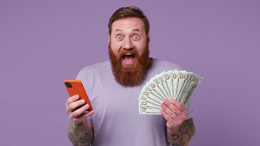 Excited happy joyful exultant young man 30s wear casual clothes use mobile cell phone hold win fan of cash money in dollar banknotes isolated on plain pastel light purple background studio portrait