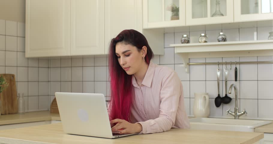 Young woman with pink hair, using laptop computer in the kitchen. Portrait of business woman watching news on laptop screen and working or studing at home workplace. Home office concept.